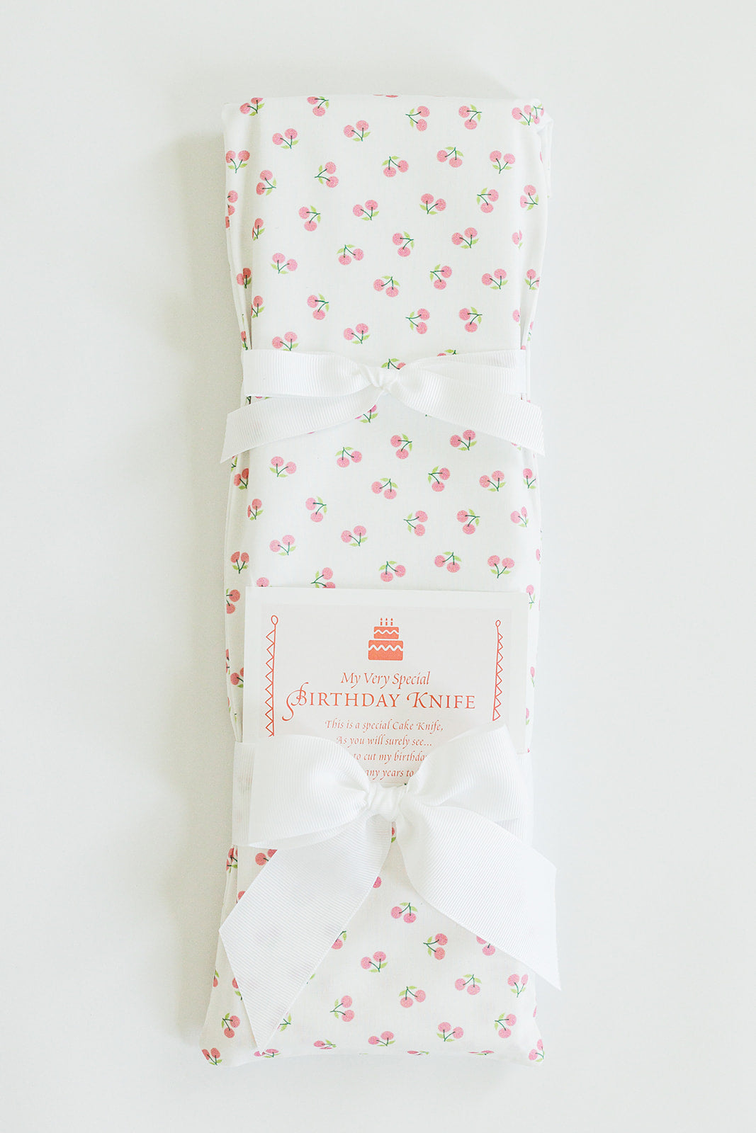A folded, white cloth with a pink cherry pattern is tied with a white ribbon. A label reads, "Jubilee Keepsakes Cherries" and describes it as the perfect cake-cutting utensil for your next birthday celebration.
