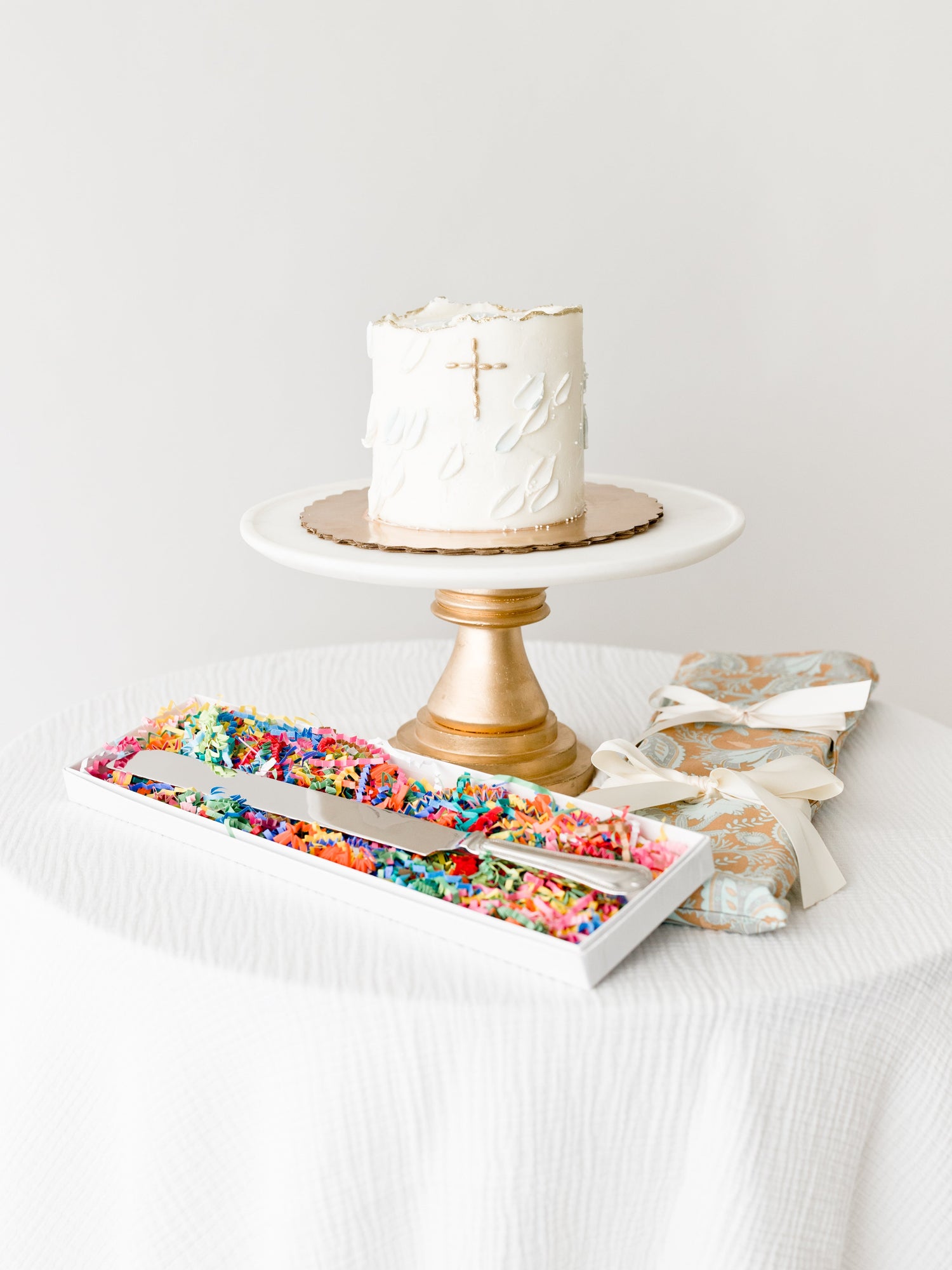 A white cake with a cross on a wooden stand sits on a table, accompanied by a colorful candy box and a wrapped gift.