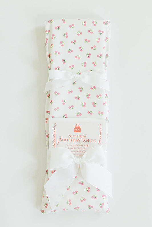 A folded, white cloth with a pink cherry pattern is tied with a white ribbon. A label reads, "Jubilee Keepsakes Cherries" and describes it as the perfect cake-cutting utensil for your next birthday celebration.