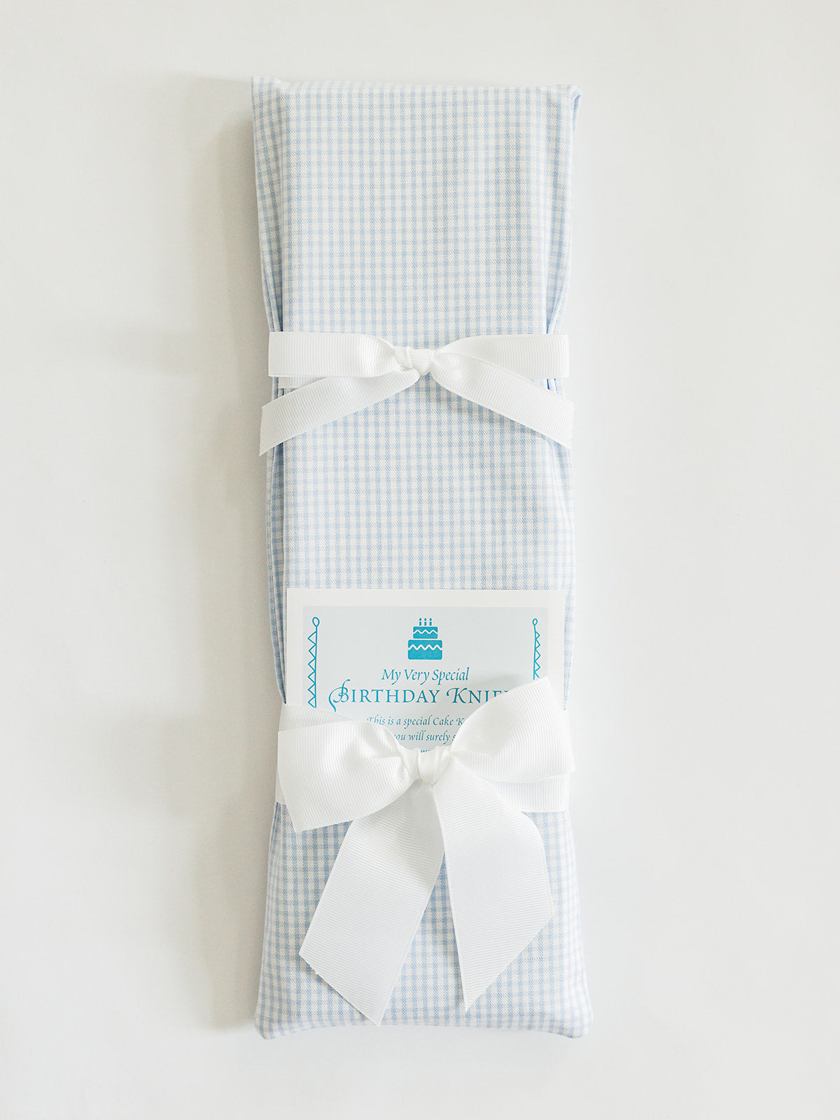 A rectangular gift wrapped in Jubilee Keepsakes' Tiny Blue Gingham paper with two white bows, a "Birthday Knit" card, and a Birthday Knife from the Birthday Knives Collection tucked beside it.