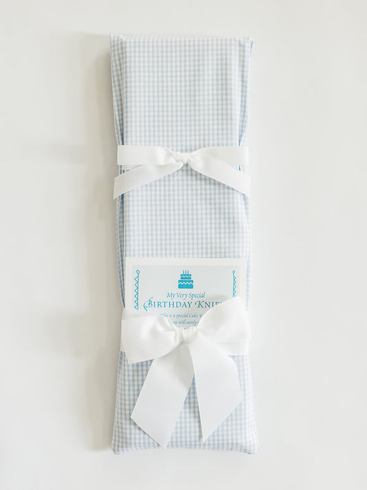 A rectangular gift wrapped in Jubilee Keepsakes' Tiny Blue Gingham paper with two white bows, a "Birthday Knit" card, and a Birthday Knife from the Birthday Knives Collection tucked beside it.